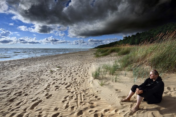 Woman sitting relaxing on sandy beach under dramatic sky, Vitrupe, Latvia