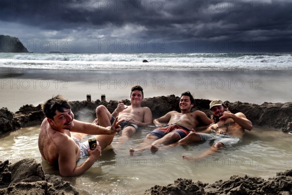 Group of men relaxing in waterhole on beach under dramatic clouds, Hot Water Beach, Coromandel Peninsula, New Zealand
