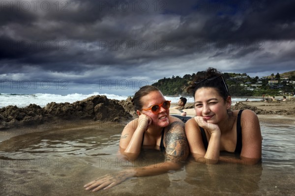 Two woman lying relaxing in a puddle of water on a beach under cloudy sky, Hot Water Beach, Coromandel Peninsula, New Zealand