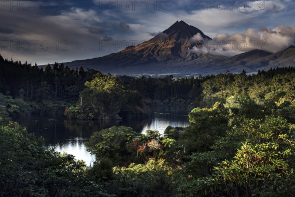 Mount Taranaki rises majestically above Lake Mangamahoe surrounded by lush greenery, New Zealand, zero