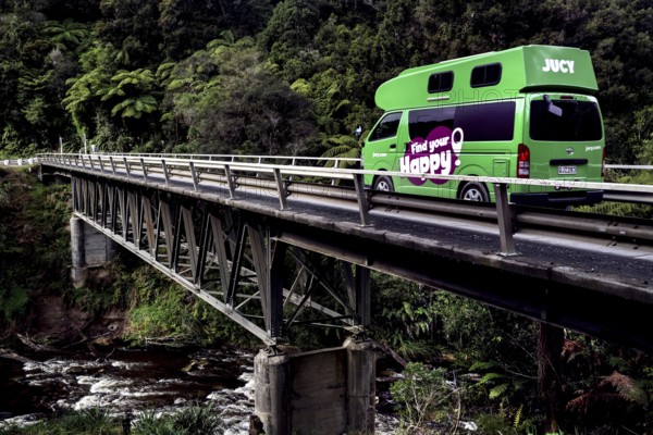 A Jucy camper crosses a bridge across the Tangarakau River on the Forgotten World Highway, surrounded by thick forest, zero
