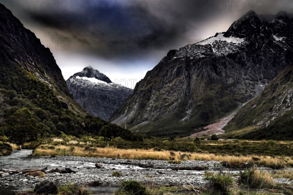 Dramatic landscape with snow-capped mountains and a river in the foreground under a cloudy sky, Milford Road, New Zealand