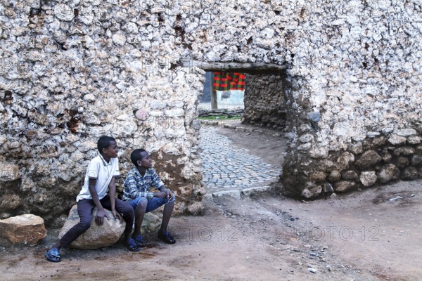 Two boys sit in front of a hyena gate in an old stone wall in the old town of Harar, Harar, Harar, Ethiopia
