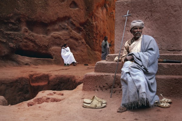 A priest with a cross sits near the rock church of Bete Emanuel in Lalibela, Lalibela, Amhara, Ethiopia