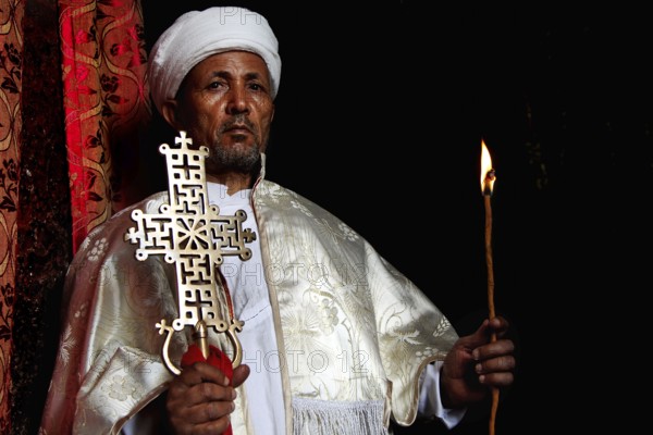 Priest in white cloak majestically highlights a cross in the dark interior of Bete Meskel, Lalibela, Amhara, Ethiopia