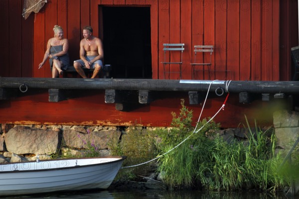 Couple sitting at a red warehouse on the shore with a boat at the water, Porvoo, Uusimaa, Finland