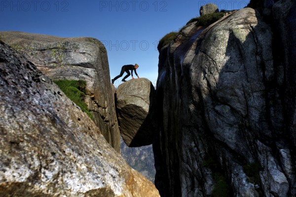 Tourist balancing on Kjeragbolten between imposing cliffs under blue sky, Lysefjord, Rogaland, Norway