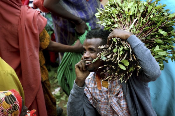 Smiling man carrying khat bundles on his shoulder and talking on the phone at a market, Awaday, Afar, Ethiopia
