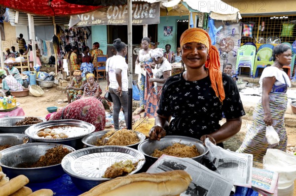 Woman in a busy market smiling behind a table with various foods, Brobo, Côte d'Ivoire