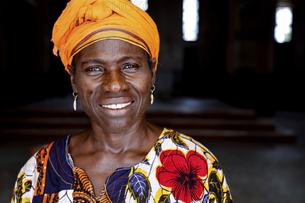 Woman in colorful dress with yellow headscarf smiles at the camera. Dark background, Bouaké, Centre, Côte d'Ivoire