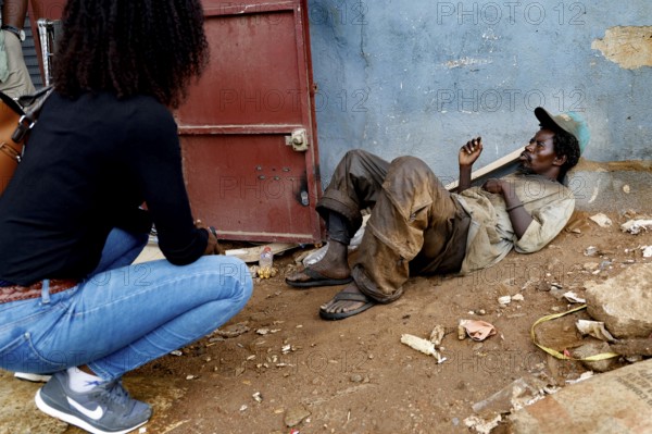 Mentally ill homeless person sits in front of a door. A woman approaches him on a dusty road, Bouaké, Côte d'Ivoire