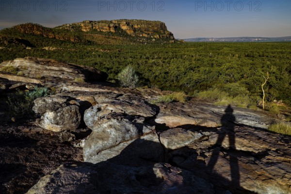 Shadows of a person on a rocky elevation in front of forest and rocks under clear sky, Nawurlandia, Northern Territory, Australia
