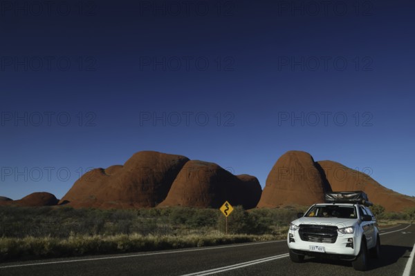 A car drives against the impressive backdrop of the Olgas under clear skies