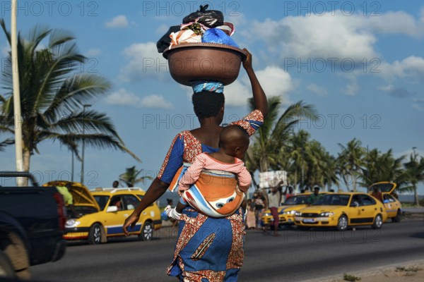 Woman carrying child on her back as palm trees and yellow taxis line the road, Abidjan, Ivory Coast