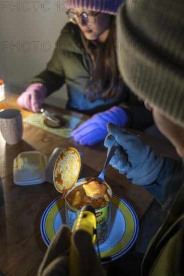 Symbolic blackout, extensive, prolonged power failure, in private surroundings, without electricity, heating, water, family at the dining table with warm winter clothes, sleeping bag, light from battery lights, flashlights, canned food, ready meals, heated on the camping stove, limited supply of food