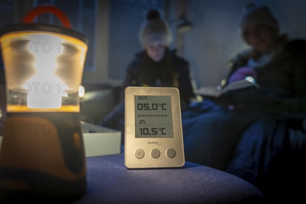 Symbolic picture blackout, extensive, prolonged power failure, in private surroundings, thermometer shows low room temperature, outside temperature, without electricity, heating, water, family on the sofa with warm winter clothes, sleeping bag, light from battery lights, battery lights, flashlights