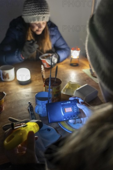 Symbolic blackout, extensive, prolonged power failure, in private surroundings, without electricity, heating, water, family at the dining table with warm winter clothes, sleeping bag, battery-operated radio, only source of information, no TV, Internet, mobile phone, light from battery lights, flashlights, food from cans, ready meals, heated on the camping stove, limited supply of food