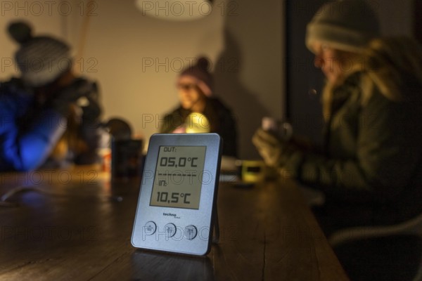 Symbolic blackout, extensive, prolonged power failure, in private surroundings, without electricity, heating, water, family at the dining table with warm winter clothes, sleeping bag, thermometer shows low room temperature, outdoor temperature, light from battery lights, flashlights, canned food, ready meals, heated on the camping stove, limited supply of food