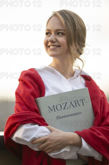 Smiling young woman, pianist, in white blouse and red cardigan, in the evening light, holding notes with Mozart, piano sonatas, in her arms, Stuttgart, Baden-Württemberg, Germany