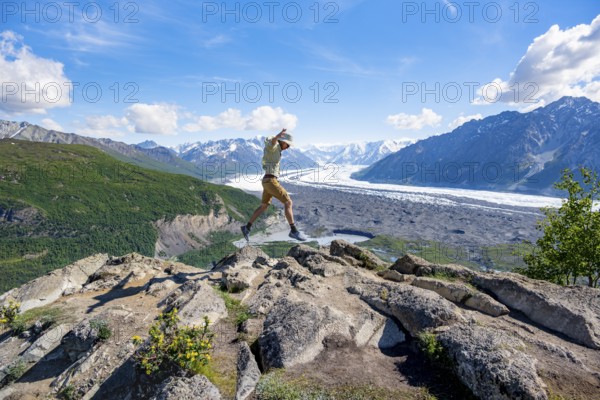 Young man jumping into the air, view of impressive mountain landscape with Matanuska glacier and glaciated mountain peaks, Lion's Head, Chugach Mountains, Alaska, USA