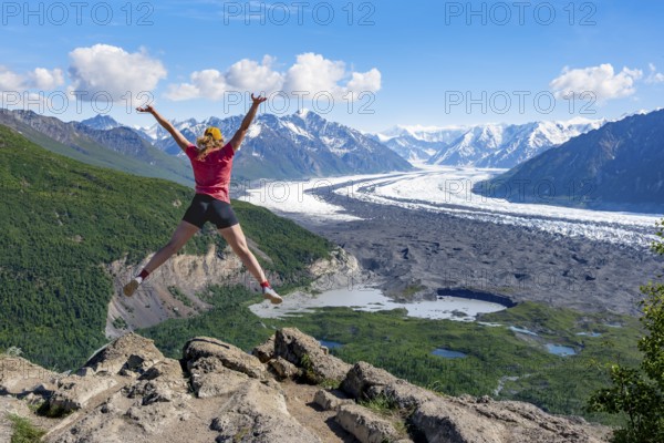 Young woman jumping into the air, view of impressive mountain landscape with Matanuska glacier and glaciated mountain peaks, Lion's Head, Chugach Mountains, Alaska, USA