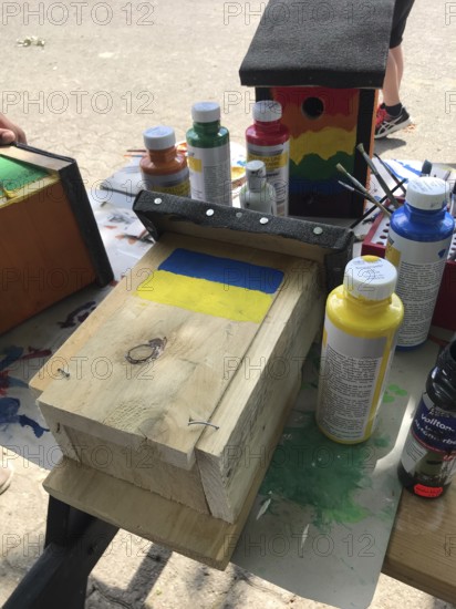 Lower Saxony, Germany, Unpainted birdhouse and brush with Ukrainian flag on a table with paints and painting supplies, painted birdhouse in blue and yellow with flowers. Ukrainian students' creative craft project with spring-like design
