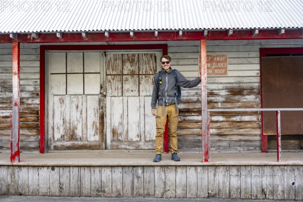 Young man standing on the porch of a former Gold Mine Independence Mine building in mountainous landscape, Independence Mine State Historical Park, Hatcher Pass, Talkeetna Mountains, Alaska, USA