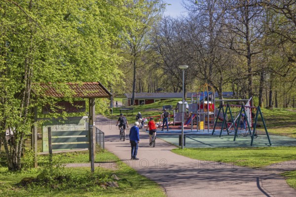 People cycling and walking by a playground with swings on a sunny spring day in a public park, Falköping, Sweden
