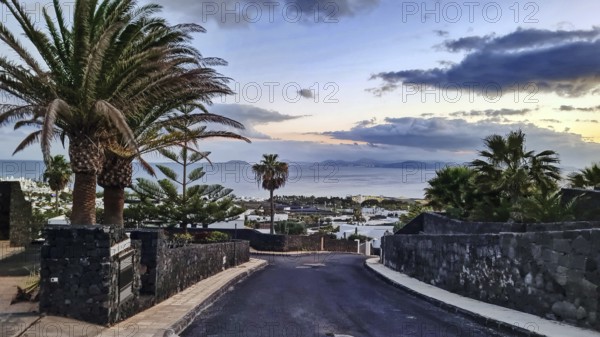 View of a road with palm trees leading to the sea, in the background the island of Fuerteventura at dusk, Playa Blanca, Lanzarote