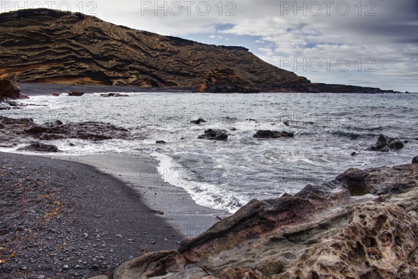 Secluded beach with black sand surrounded by wavy rock formations under a cloudy sky, Chargo Verde El Golfo Lanzarote