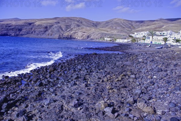 Rocky coastal landscape with blue waves and a village at the foot of the mountains under clear skies, Playa Quemada Yaiza Lanzarote