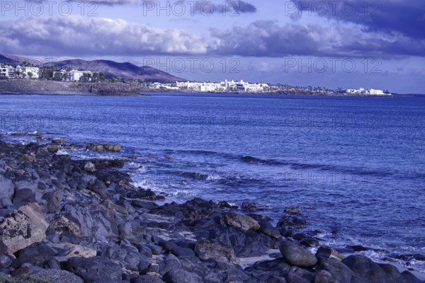 Rocky coastal landscape with views of the sea and a distant town under a cloudy sky, Playa Blanca Lanzarote