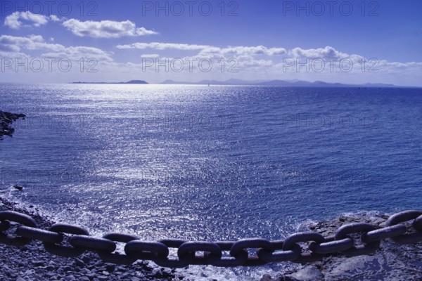 Chain on the edge of a cliff with a view of a calm, blue sea and distant island of Furteventura in the background, Playa Blanca Lanzarote