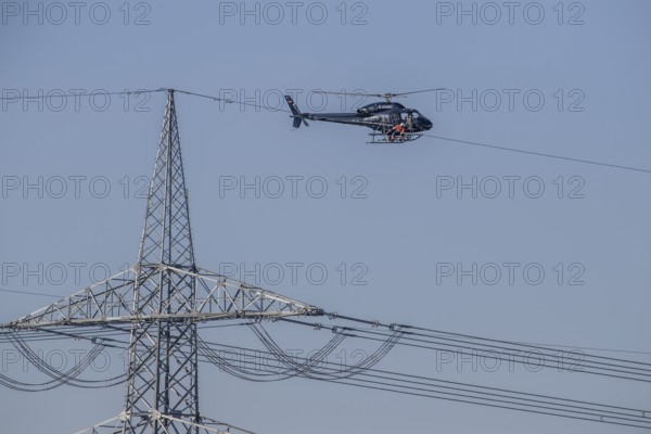 A helicopter carries out technical work on power lines next to a mast under a blue sky, an employee marks the top line of a new power line, Lemförde, Lower Saxony, Germany