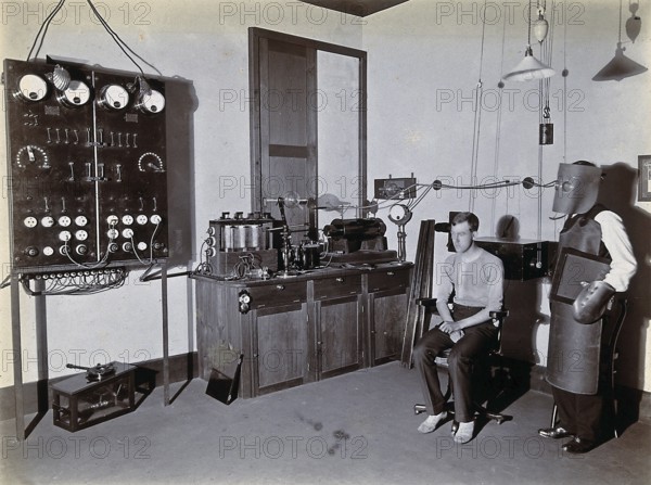 X-ray, hospital, man wearing a heavy apron, face mask and gloves to protect against X-rays, possibly with an X-ray plate in his hand next to a seated patient. Photograph, around 1905. Medicine, history of medicine, photography, Johannesburg, South Africa, historical