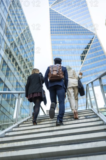 Diverse business colleagues climb a modern exterior staircase against glass skyscrapers, symbolizing teamwork, ambition and upward career progress in a dynamic urban setting