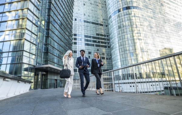 Diverse business professionals walking, communicating, and laughing while moving together in a modern financial district with glass office buildings reflecting the bright sky