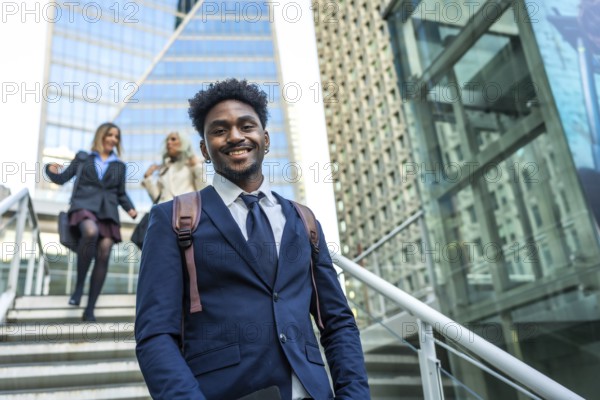 African american businessman smiling at camera and walking up stairs in a modern urban environment with female colleagues ascending steps in the background, conveying success and career growth