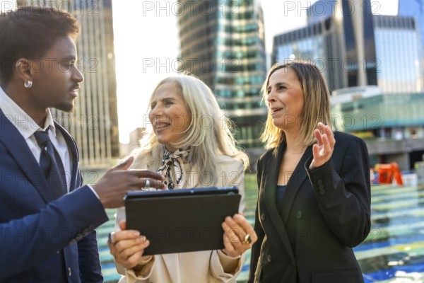 Diverse business team engaging in an informal outdoor meeting, discussing a project while looking at a digital tablet, with modern city buildings in the background