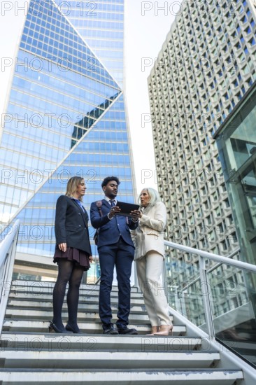 Diverse business team meeting on outdoor stairs in a downtown corporate district, collaborating over a tablet with skyscrapers behind, symbolizing teamwork and urban innovation