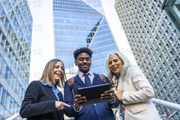 Diverse business people smiling and showing teamwork, standing outdoors in a corporate city environment while looking at a tablet together and discussing modern digital strategy