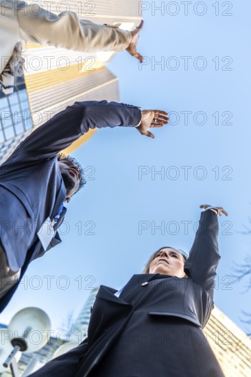 Diverse business professionals extending arms upwards, aiming for a shared objective and future growth against the backdrop of modern skyscrapers and a clear blue sky