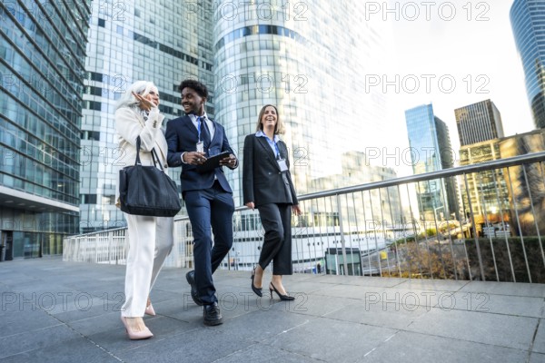 Diverse business professionals senior woman, black man, and young woman walk through a modern corporate streetscape, smiling, discussing strategy and sharing a tablet