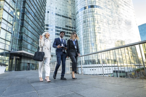 Diverse business professionals, including a senior woman, walking together on an urban walkway outdoors, holding documents and discussing work with modern glass skyscrapers in the background