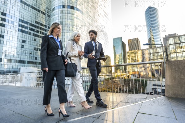 Three diverse business people walking outdoors on an elevated pedestrian area, engaged in discussion with modern corporate buildings reflecting sunlight in the background