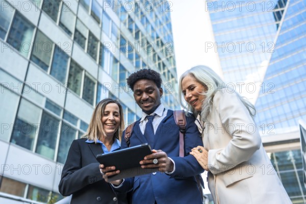 Diverse business professionals reviewing content on a digital tablet, fostering collaboration and teamwork while standing in front of modern corporate skyscrapers and discussing strategy