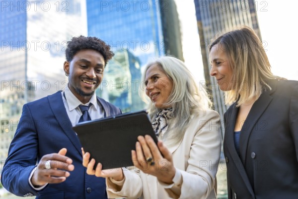 Diverse business professionals reviewing information on a digital tablet, standing outdoors in front of modern corporate buildings enjoying a collaborative meeting