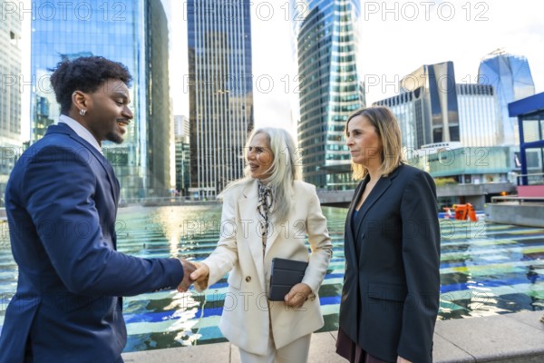 Diverse business professionals, a black man and two women, are shaking hands and discussing in a modern financial district with glass skyscrapers reflecting the light
