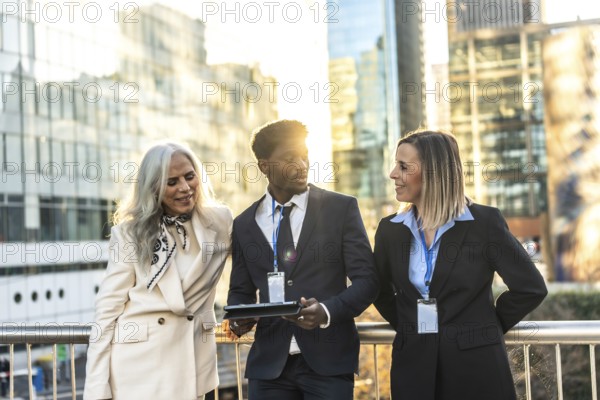 Diverse group of business people collaborating and discussing work in an urban setting, featuring an african american man holding a tablet while communicating with two female colleagues