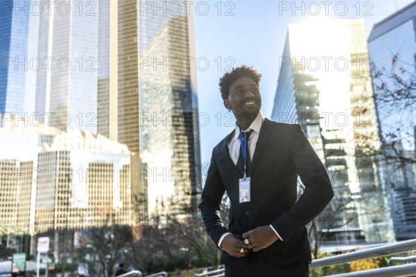 Young african american businessman in a dark suit and id badge smiles confidently in a sunny urban business district, looking toward the future amid skyscrapers and opportunity